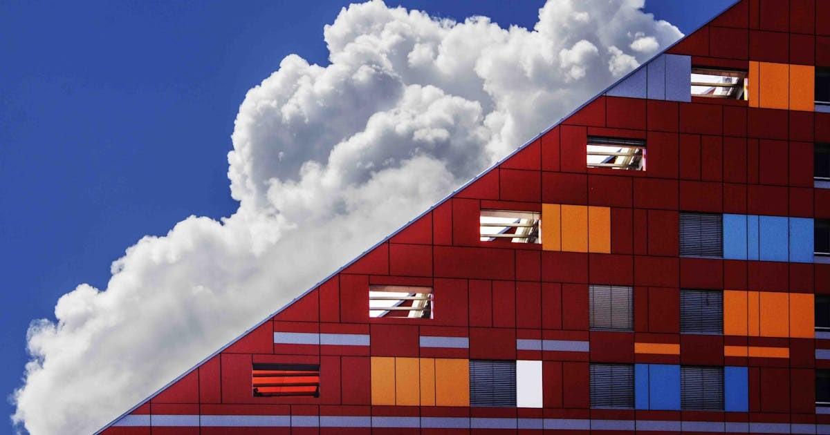 a modern building with red, orange, and blue panels with a large white cloud above it against a blue sky